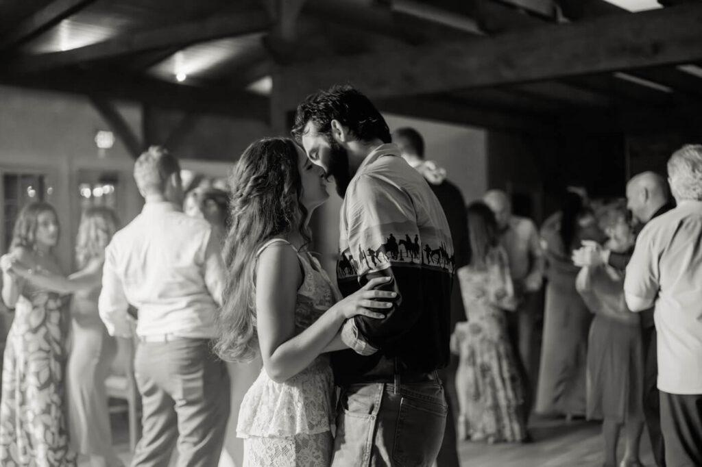 Bride and groom’s wedding dance kiss – A black-and-white image captures the newlyweds kissing on the dance floor, surrounded by guests in the warm ambiance of their barn wedding reception.