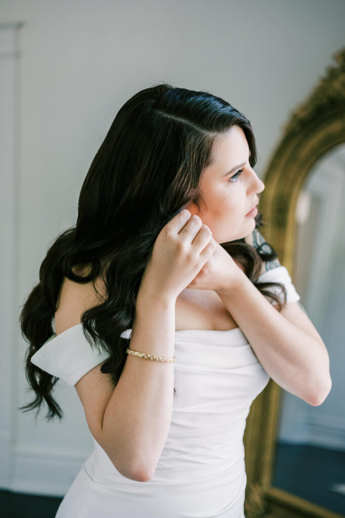 Bride Putting on Earrings Before Wedding Ceremony – A close-up of the bride with soft waves in her hair, delicately placing her earrings as natural light highlights her refined and timeless bridal look.