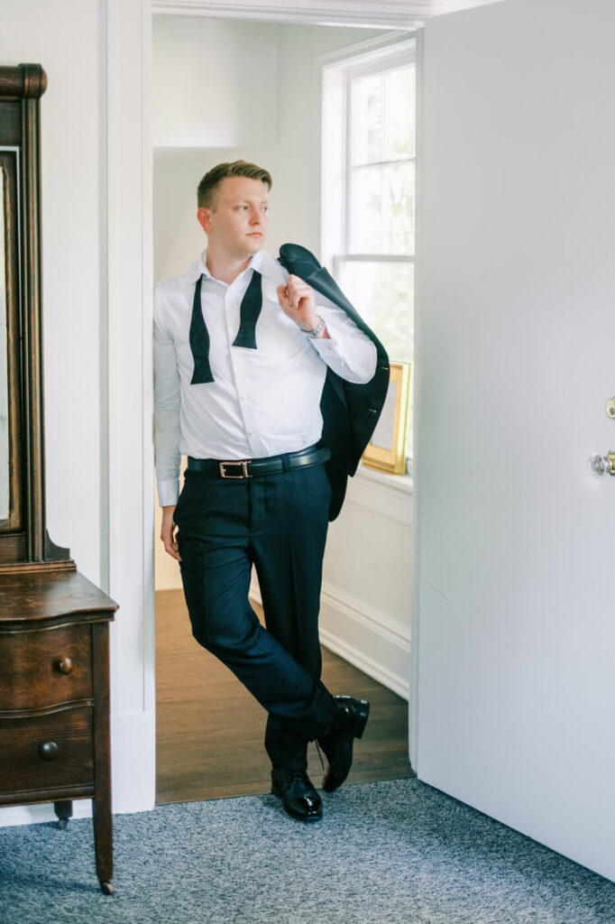 Groom Prepares for His Wedding at McAlister-Leftwich House – The groom leans against the doorway with a relaxed yet sophisticated pose, his suit jacket draped over his shoulder, capturing an effortless, classic groom’s style.