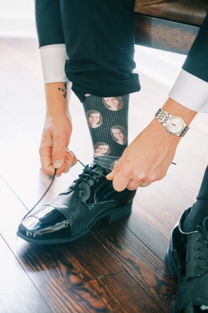 Groom Tying Shoelaces with Custom Wedding Socks – A candid moment of the groom lacing up his polished black dress shoes, revealing custom socks with the bride’s face, adding a touch of personality to the wedding morning.