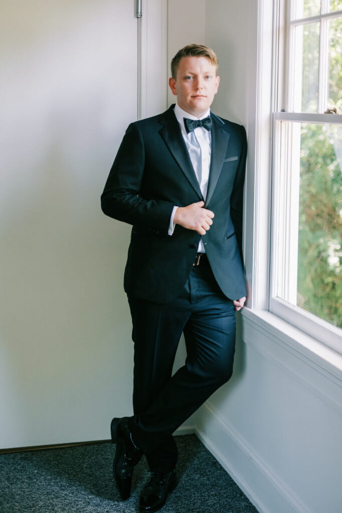 Classic Groom Portrait by the Window in Greensboro, NC – The groom stands elegantly by the window in a black tuxedo, exuding sophistication and confidence as he prepares for the wedding ceremony.