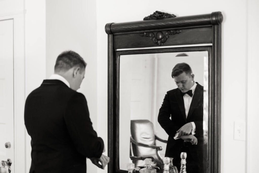 Groom Fixing Cufflinks in Front of Antique Mirror – A black-and-white image of the groom adjusting his cufflinks, reflected in an antique mirror, adding a refined, vintage charm to the wedding day moments.