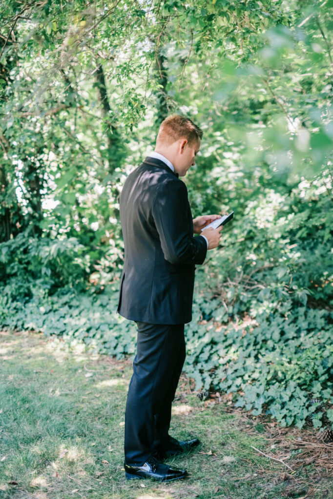 Groom reading vows in a private garden moment – A groom dressed in a classic black tuxedo reading a personal note in a lush, green garden before his wedding ceremony at McAlister-Leftwich House.