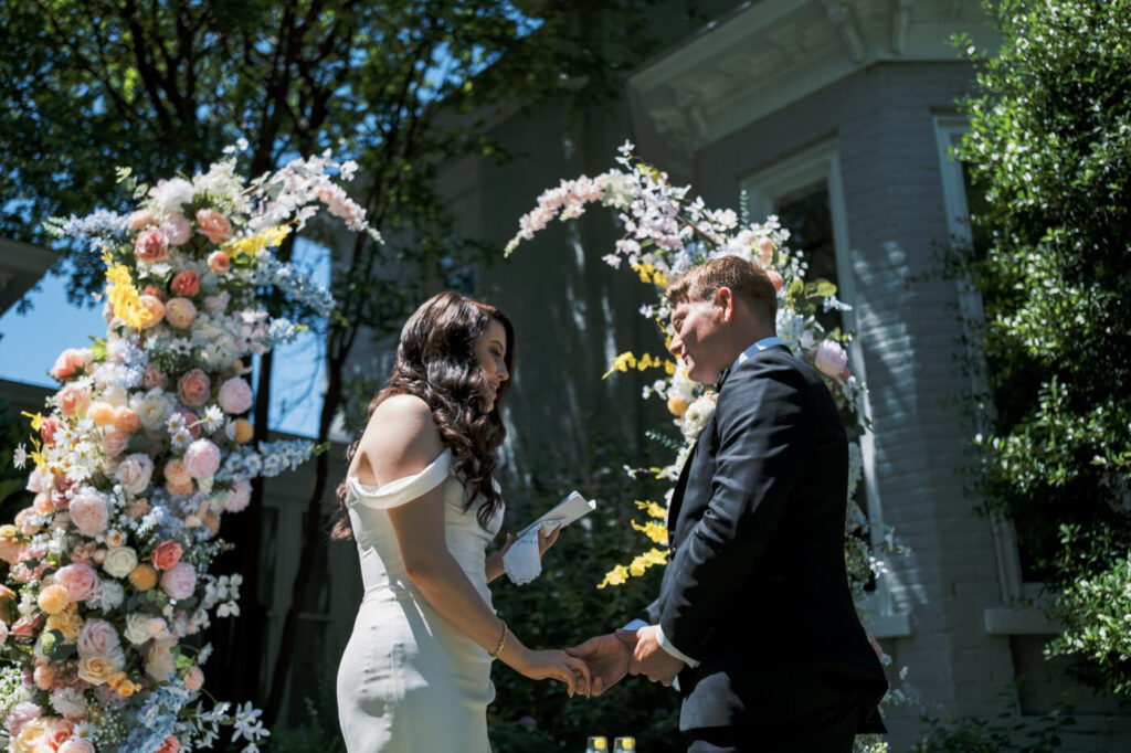 Bride and groom exchanging vows under a floral arch – A joyful bride and groom holding hands under a pastel floral arch in the garden of McAlister-Leftwich House, sharing emotional vows on their wedding day.