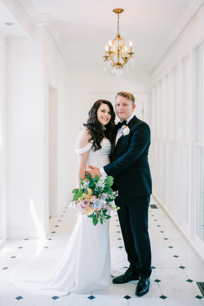 Bride and groom posing in an elegant white hallway – A radiant couple stands together in a bright, white hallway with a gold chandelier, capturing their timeless wedding day look at McAlister-Leftwich House.