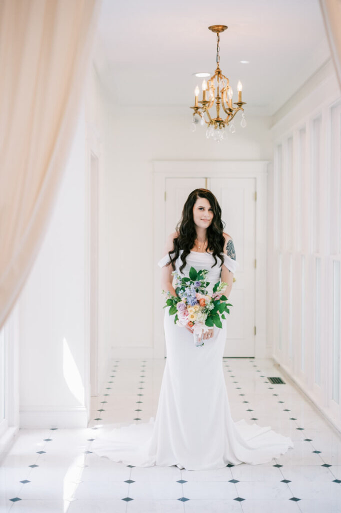 Bridal portrait in an elegant, light-filled hallway – A stunning bride in a fitted white gown holding a pastel floral bouquet, standing gracefully in a white hallway at McAlister-Leftwich House.