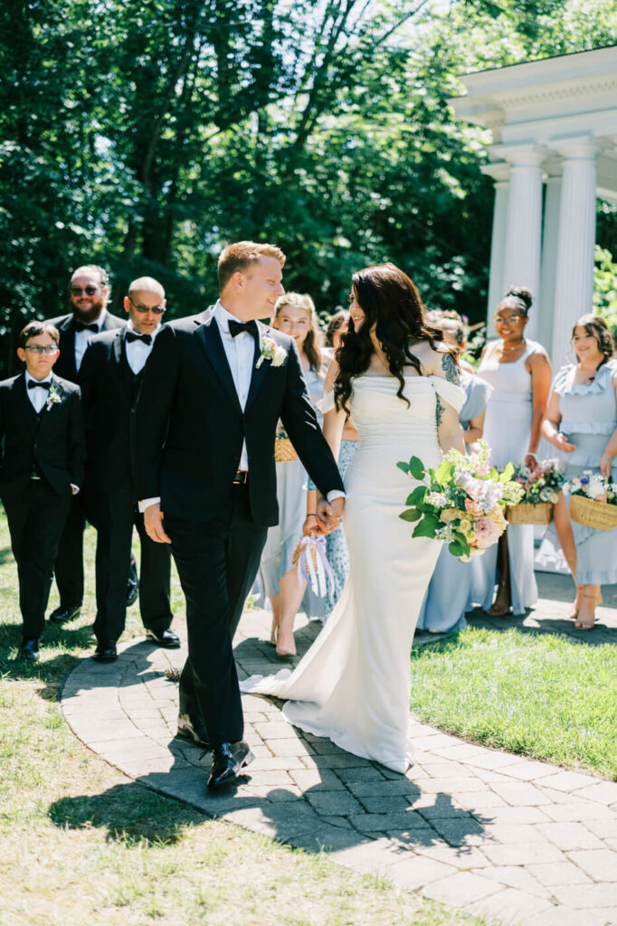 Bride and groom leading their wedding party in celebration – A newlywed couple smiling as they walk hand in hand, surrounded by a joyful wedding party in coordinated pastel blue attire at McAlister-Leftwich House.