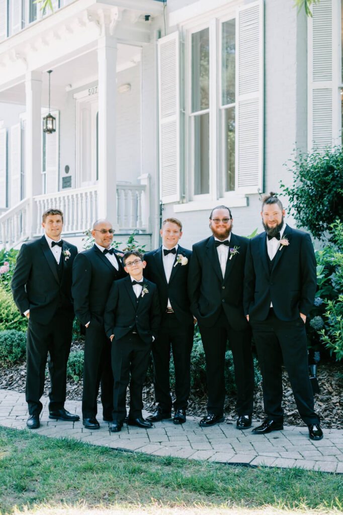 Groom and groomsmen posing in classic black tuxedos – A groom and his groomsmen, dressed in sophisticated black tuxedos, pose in front of the historic McAlister-Leftwich House, exuding timeless wedding elegance.