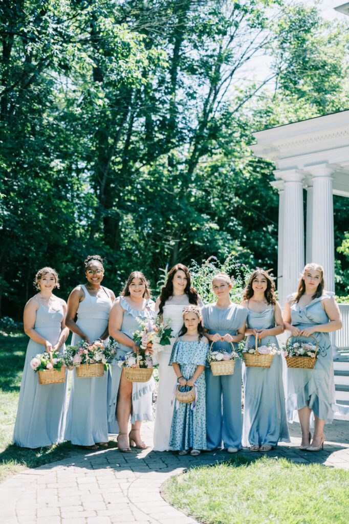 Bridesmaids in blue dresses holding floral baskets – A bride and her bridesmaids in flowing blue dresses hold charming floral baskets, standing in a sunlit garden at McAlister-Leftwich House.