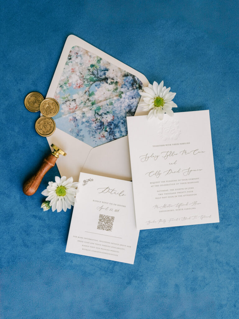 Wedding Invitation Flat Lay – Elegant wedding invitation suite with a floral envelope liner, gold wax seal, and delicate daisy flowers, styled on a deep blue background.