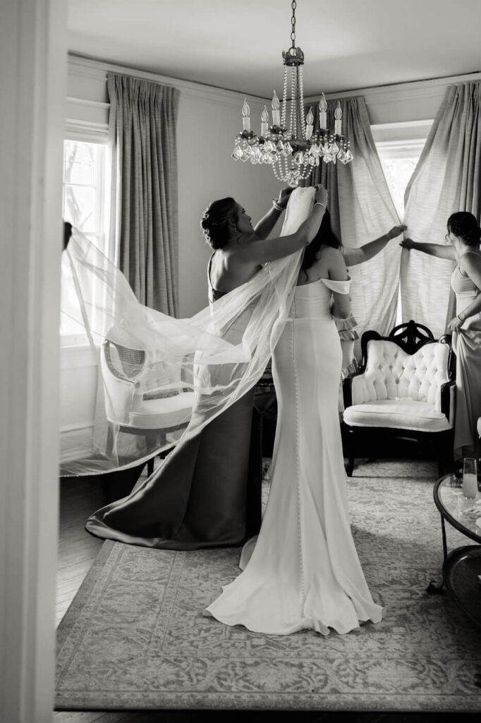 Bridal Veil Placement in Historic Wedding Venue – A timeless black-and-white photograph of the bride’s attendants adjusting her long flowing veil in a beautifully decorated McAlister-Leftwich House bridal suite with a chandelier.