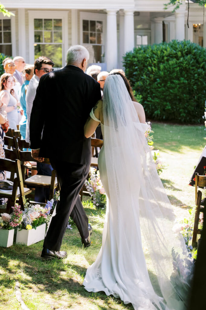 Bride Walking Down the Aisle at McAlister-Leftwich House – The bride walks arm-in-arm with her father down the outdoor ceremony aisle, her veil trailing behind as guests watch with admiration.