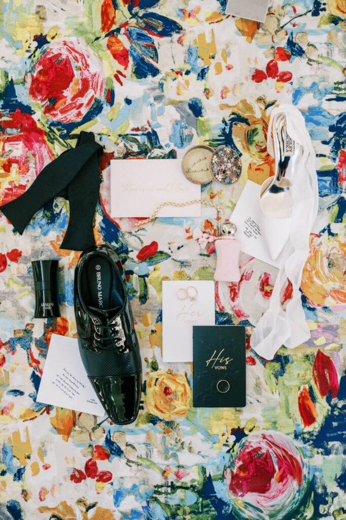 Bridal and Groom Wedding Details – Artistic flat lay of bride and groom wedding accessories, including vow books, a bow tie, jewelry, shoes, and luxury cologne, placed on a colorful floral backdrop.