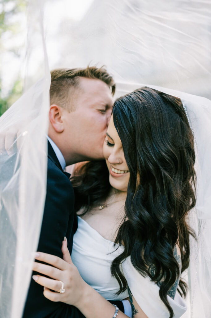 Romantic Wedding Veil Portrait of Bride and Groom – The groom gently kisses his bride’s forehead under the soft, flowing veil, capturing an intimate and timeless moment on their wedding day.