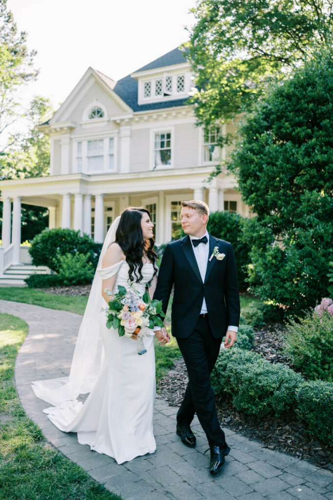 Bride and Groom Walking at McAlister-Leftwich House – A newlywed couple walks hand in hand on a garden path in front of the historic McAlister-Leftwich House in Greensboro, NC, surrounded by lush greenery.