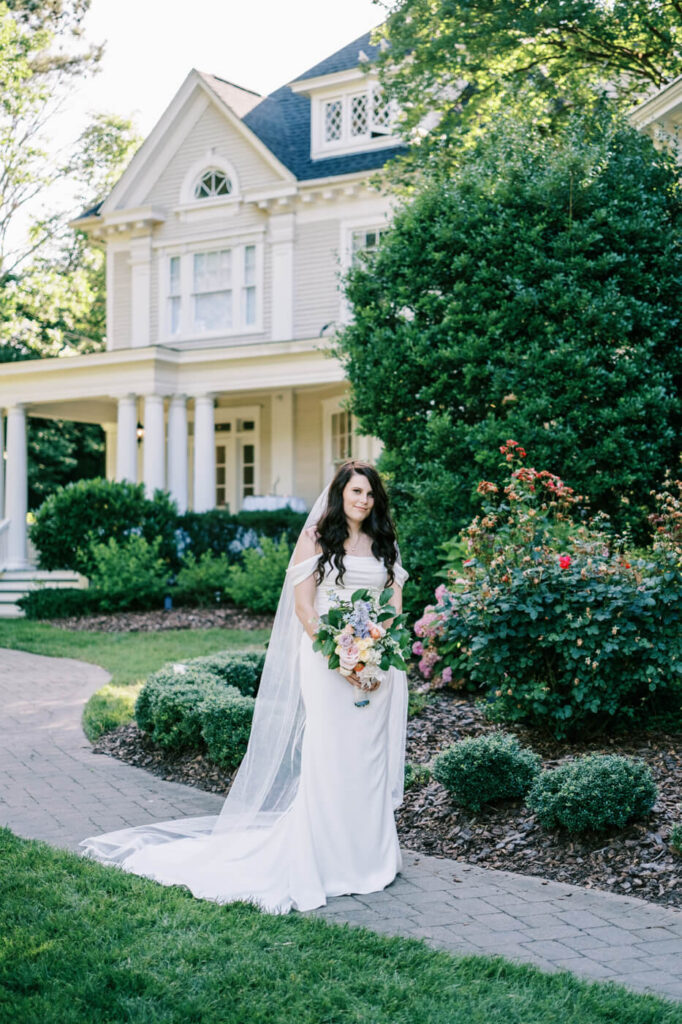 Bride Holding Bouquet in Front of McAlister-Leftwich House – A bride in a white gown stands gracefully in a garden, holding a pastel floral bouquet in front of the McAlister-Leftwich House in Greensboro, NC.