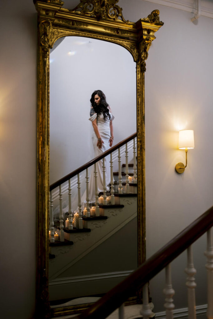 Bride Reflected in Ornate Gold Mirror on Staircase – A bride in a sleek white wedding dress is reflected in a grand antique mirror, surrounded by romantic candlelight on the staircase.