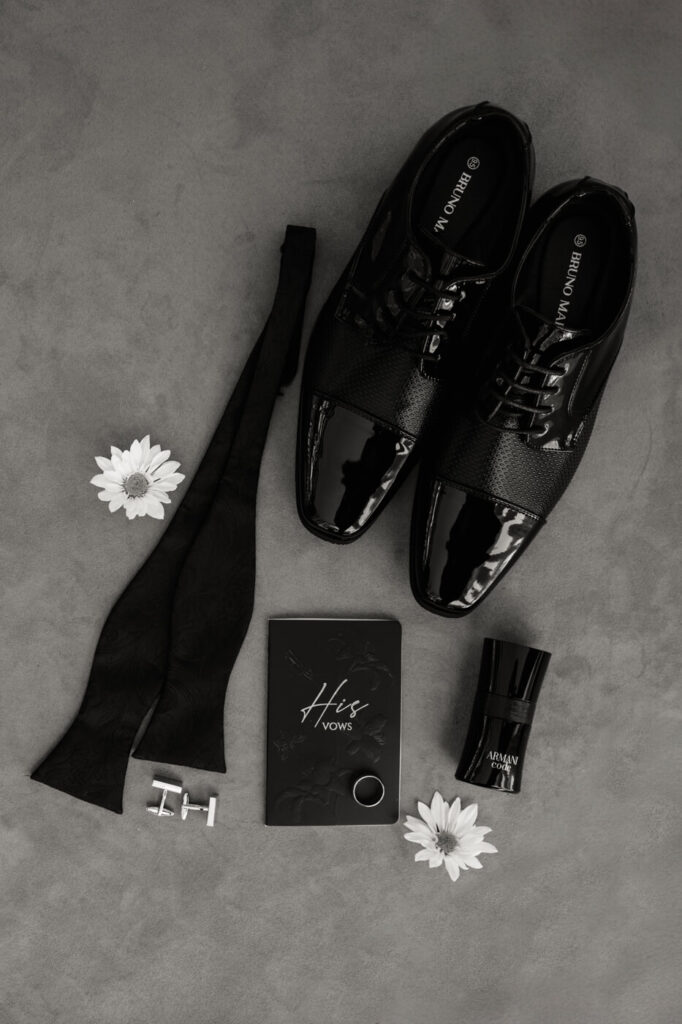 Groom’s Classic Black Tie Accessories – A black and white groom’s detail shot featuring patent leather shoes, cufflinks, a bow tie, and a personalized vow book.