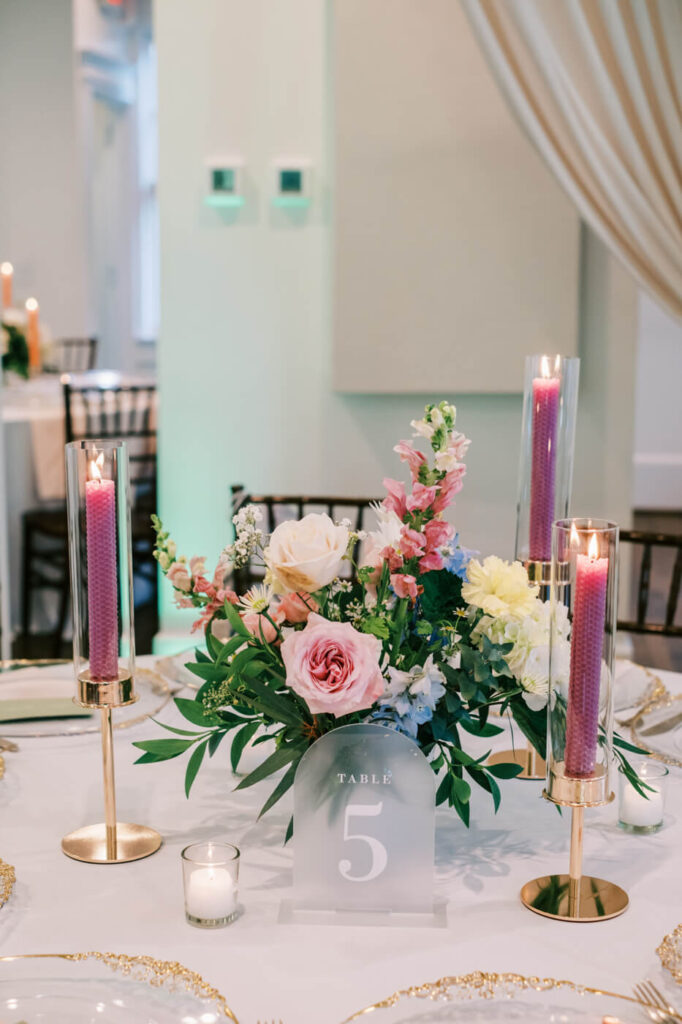 Romantic Wedding Table Centerpiece with Floral Arrangements and Candles – A close-up of a wedding reception table adorned with pastel florals, tall glass candles, and an elegant frosted table number.