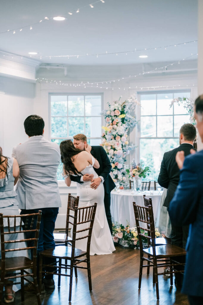 Just married couple shares first kiss at McAlister-Leftwich House – A groom embraces his bride for a heartfelt kiss during their wedding ceremony, framed by soft floral arrangements and glowing natural light.