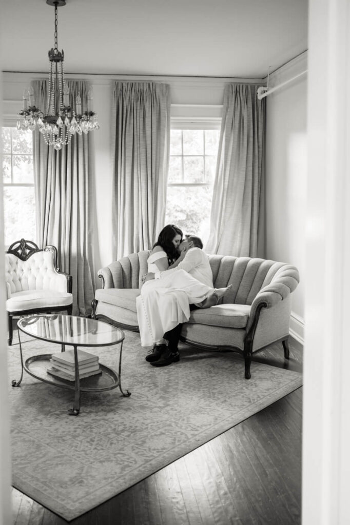 Candid newlywed moment on an elegant vintage sofa – A black-and-white photograph of a newly married couple cuddling on a classic tufted couch, surrounded by vintage decor and soft drapery.