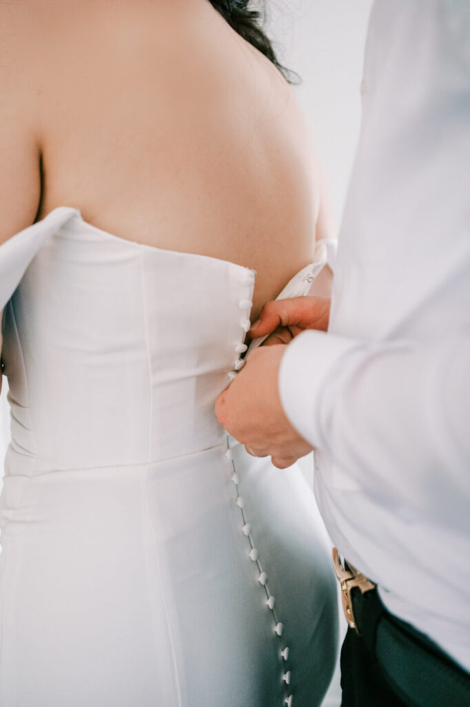 Bride’s wedding dress being buttoned up by her groom – A close-up of the groom fastening the delicate buttons on the back of the bride’s classic white gown, a beautiful pre-ceremony moment.