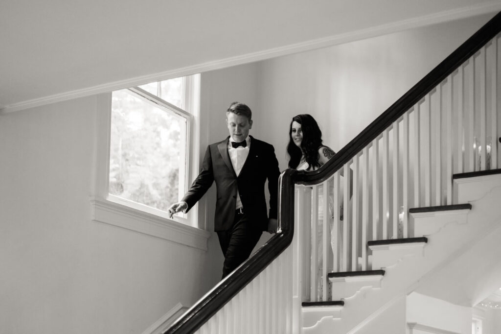 Bride and groom walking down the historic staircase of McAlister-Leftwich House – A black-and-white photo capturing the couple’s elegant entrance down a grand staircase, a timeless Greensboro wedding venue.