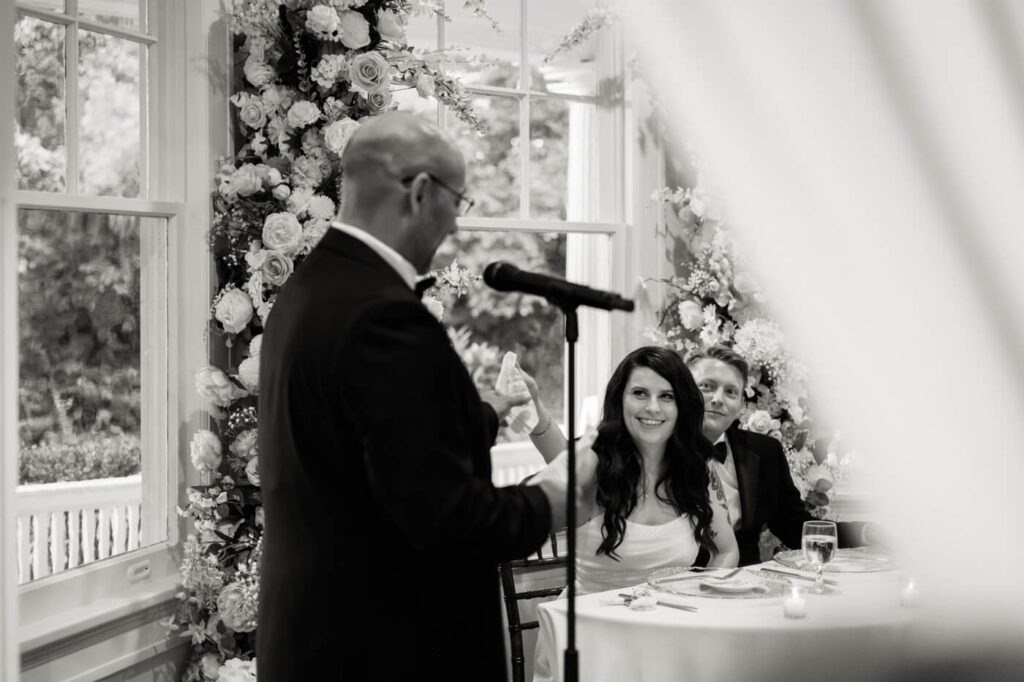 Heartfelt wedding toast at McAlister-Leftwich House reception – The bride smiles as she listens to a speech, framed by lush floral arrangements and the soft glow of the historic venue’s windows.