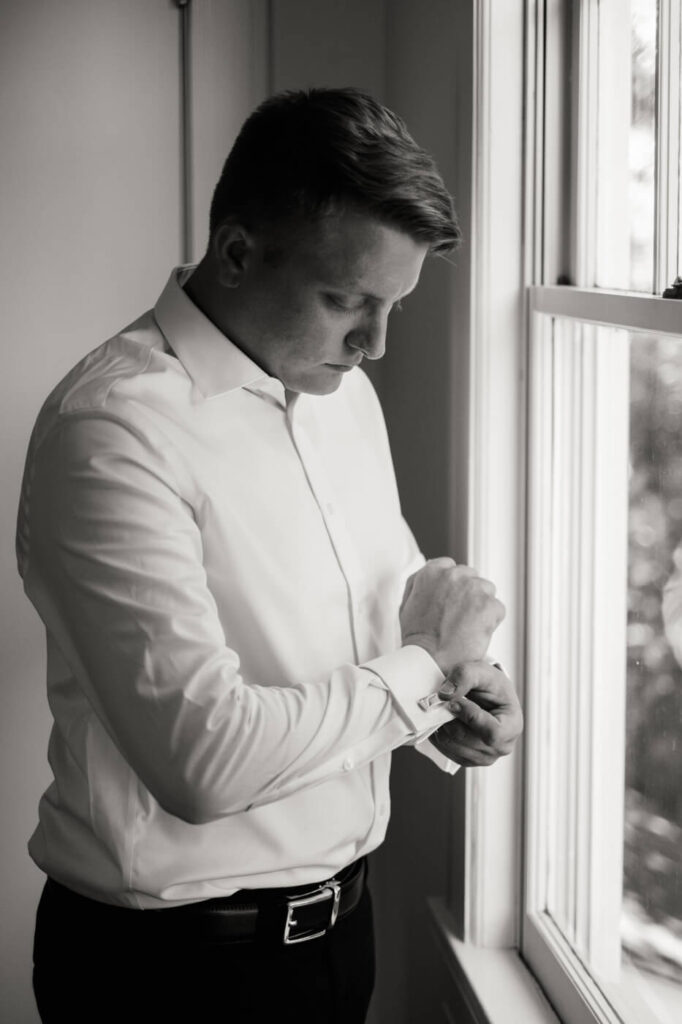 Groom Reflection Portrait – A moody black and white image of the groom sitting in an antique chair, adjusting his shirt sleeves before the ceremony.