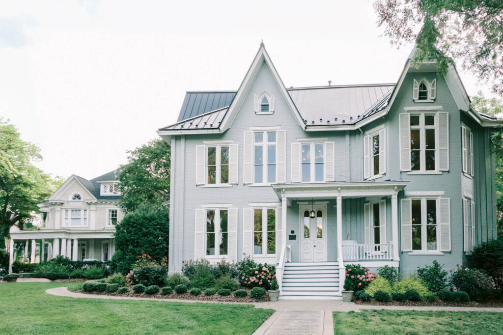 The charming exterior of McAlister-Leftwich House in Greensboro, NC – A stunning historic mansion with soft blue-gray walls, white shutters, and lush gardens, the perfect venue for a timeless Southern wedding.