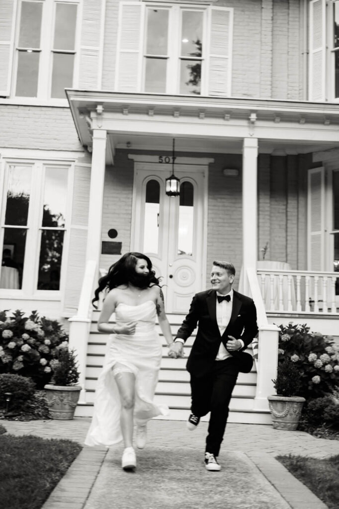 Black and white photo of a newlywed couple running hand in hand in front of McAlister-Leftwich House, showcasing their playful and joyful wedding celebration.