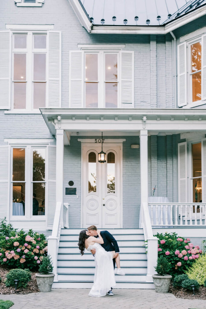 Groom dips and kisses his bride on the grand steps of McAlister-Leftwich House, surrounded by lush greenery and classic Southern charm.