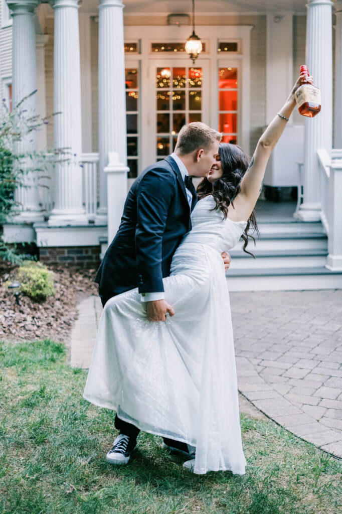 Newlyweds share a celebratory kiss in front of a historic estate, with the bride raising a bottle of bourbon in pure wedding-day bliss.