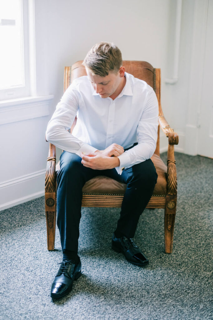 Groom Getting Ready at McAlister-Leftwich House – A groom adjusts his cufflinks sitting in a chair near a window, dressed in a crisp white shirt, preparing for the wedding day.