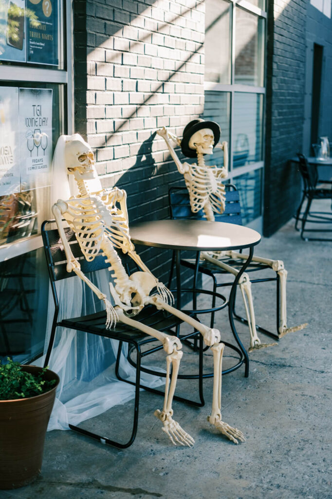 Outdoor skeleton couple wedding decor – A whimsical touch to the wedding rehearsal dinner décor featuring a skeleton bride in a veil and a skeleton groom in a hat seated at a café table.