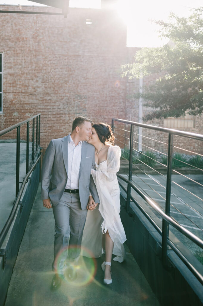 Sunlit moment between the bride and groom as they walk hand in hand on a modern industrial-style bridge during their rehearsal dinner.