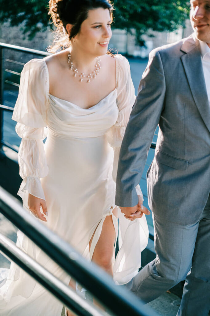 Bride and groom ascend an outdoor staircase, glowing in the golden-hour light, with the bride’s puff-sleeved gown flowing behind her.