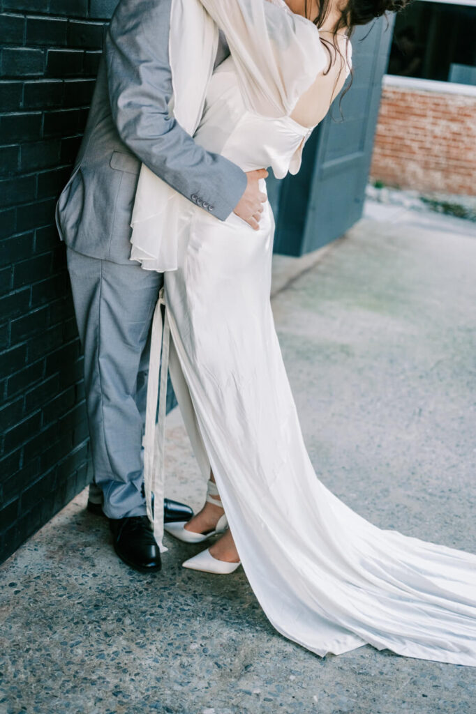 A romantic embrace between the couple against a dark brick wall, the bride’s silk gown pooling elegantly at her feet.