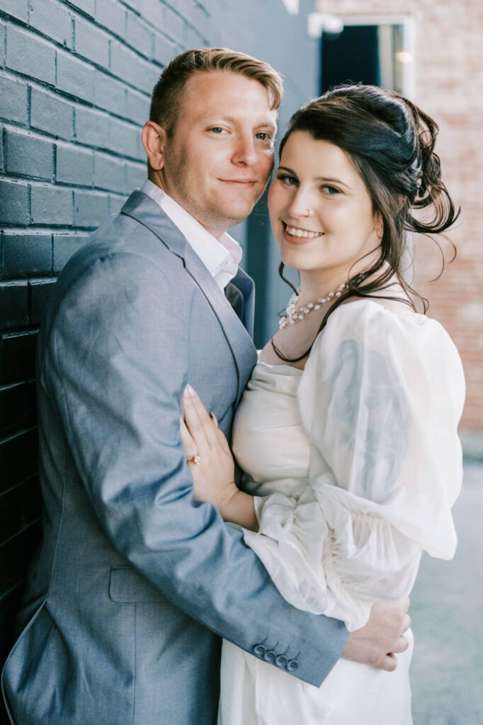 Softly smiling bride leaning into her groom, both dressed in timeless attire, against the backdrop of a historic Greensboro wedding venue.