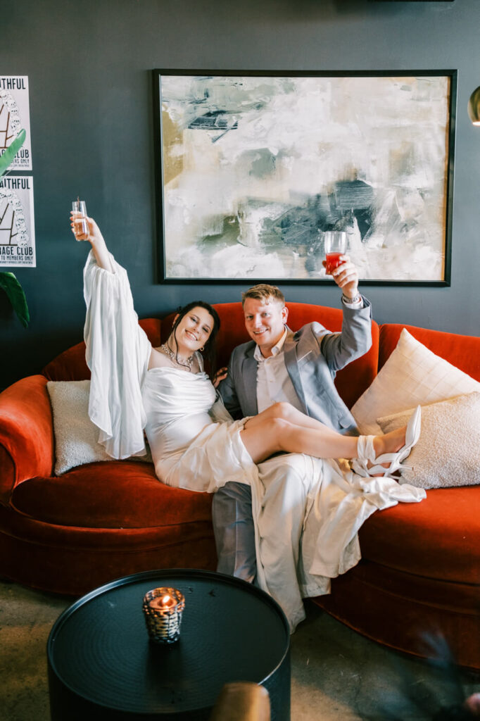 Bride and groom sharing a toast on a vintage red couch – A joyful moment as the couple raises their glasses, celebrating their upcoming wedding on a stylish velvet red couch.