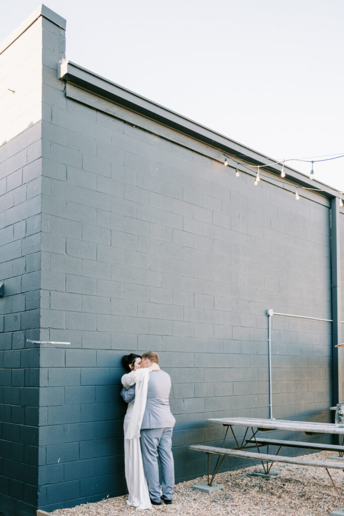 Bride and groom embracing in an industrial setting – A modern and intimate moment as the couple leans into each other against an urban brick wall, with soft evening light surrounding them.