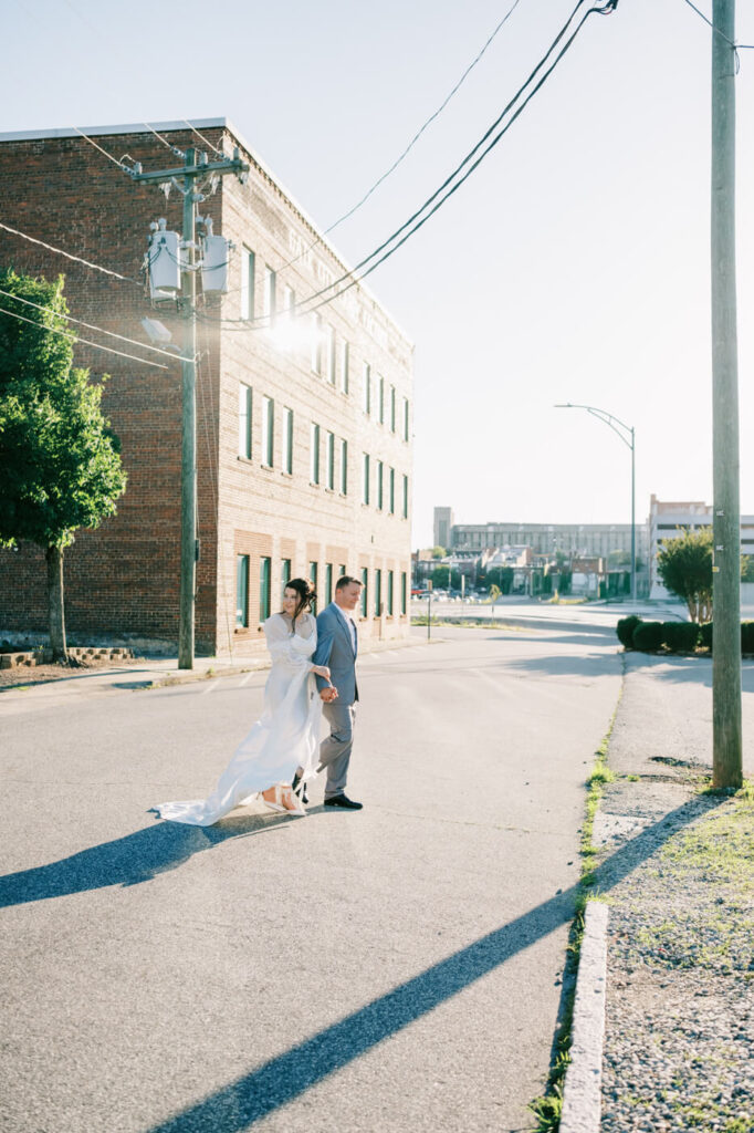 Couple walking through downtown Greensboro – The bride’s train flowing as the couple walks hand-in-hand down a city street, with the golden setting sun casting long shadows.