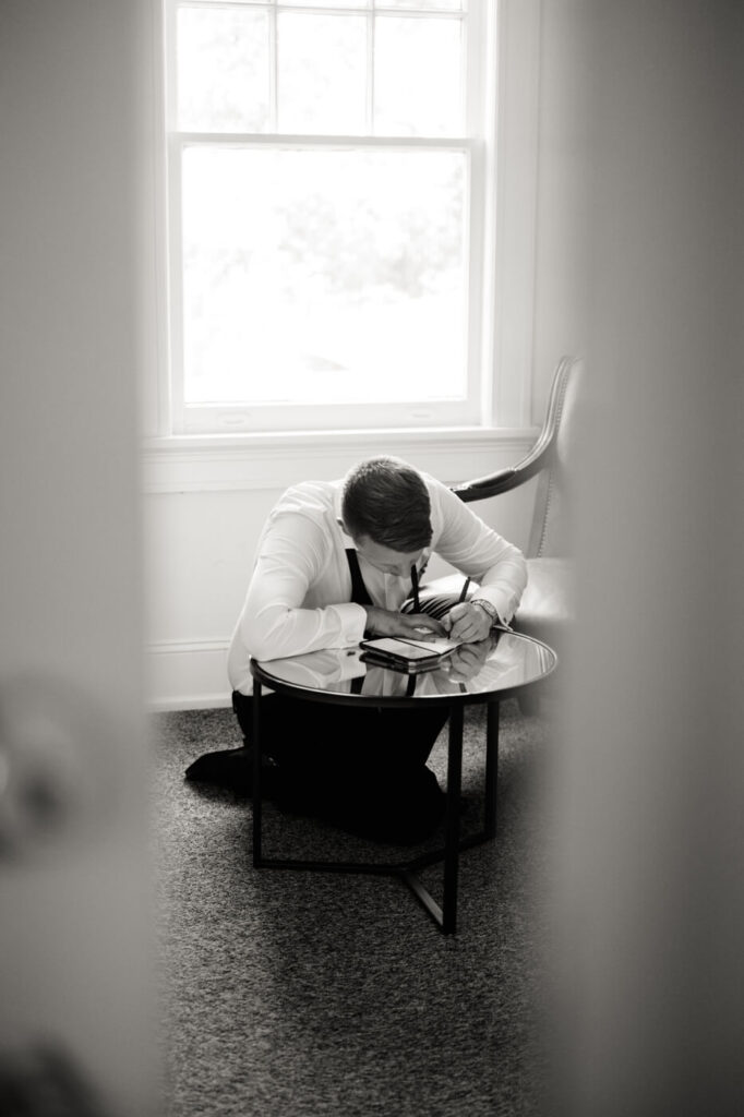 Groom Writing His Wedding Vows – Emotional black and white moment of the groom kneeling at a table, carefully writing his vows in a quiet room at McAlister-Leftwich House.