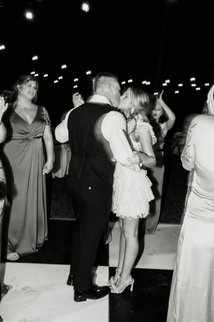 Romantic wedding dance under string lights – A bride and groom share a sweet kiss on the dance floor, surrounded by guests in elegant attire, celebrating their wedding at The Historic Robertson House.