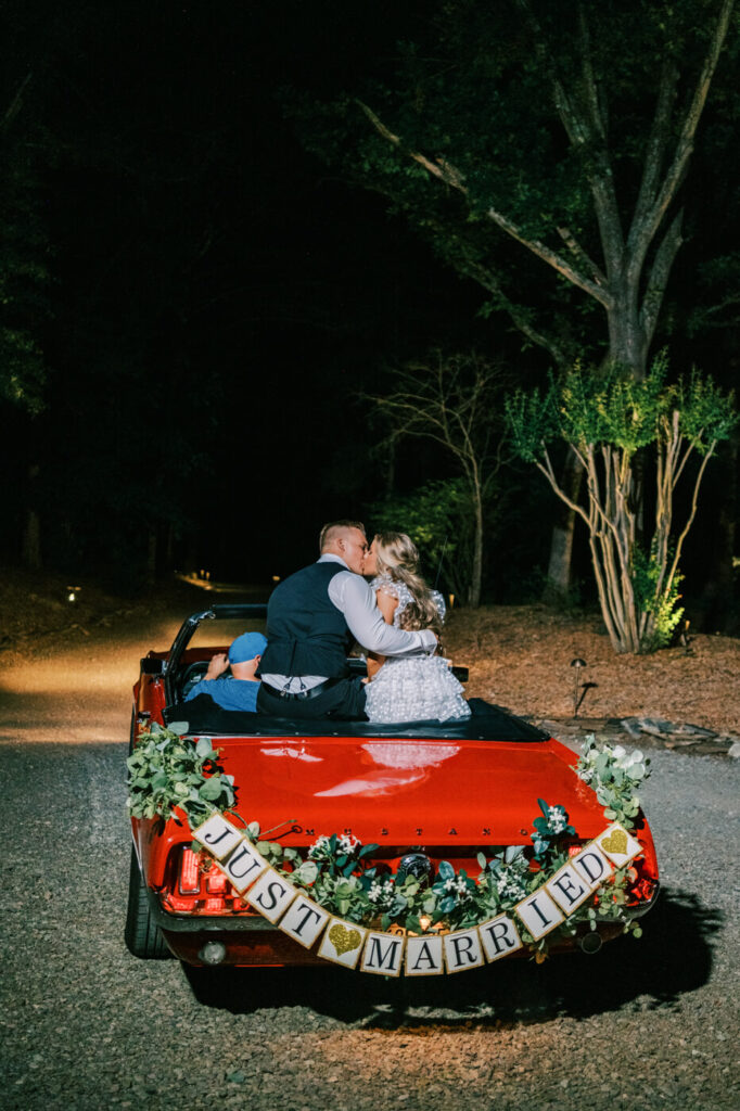 Newlyweds share a kiss in their vintage wedding getaway car – The bride and groom steal a romantic moment as they sit in their beautifully decorated red Mustang, ready to drive off into the night.