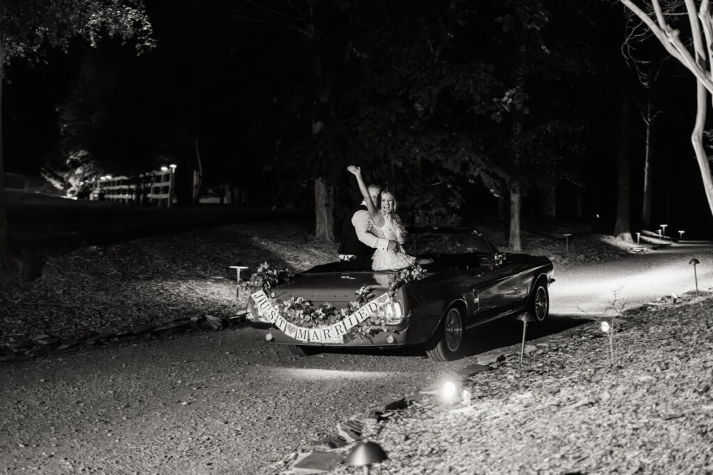 Bride waving farewell during wedding exit drive – The newlywed couple rides into the night in a vintage Mustang, with the bride happily waving to guests as they bid their final goodbyes.