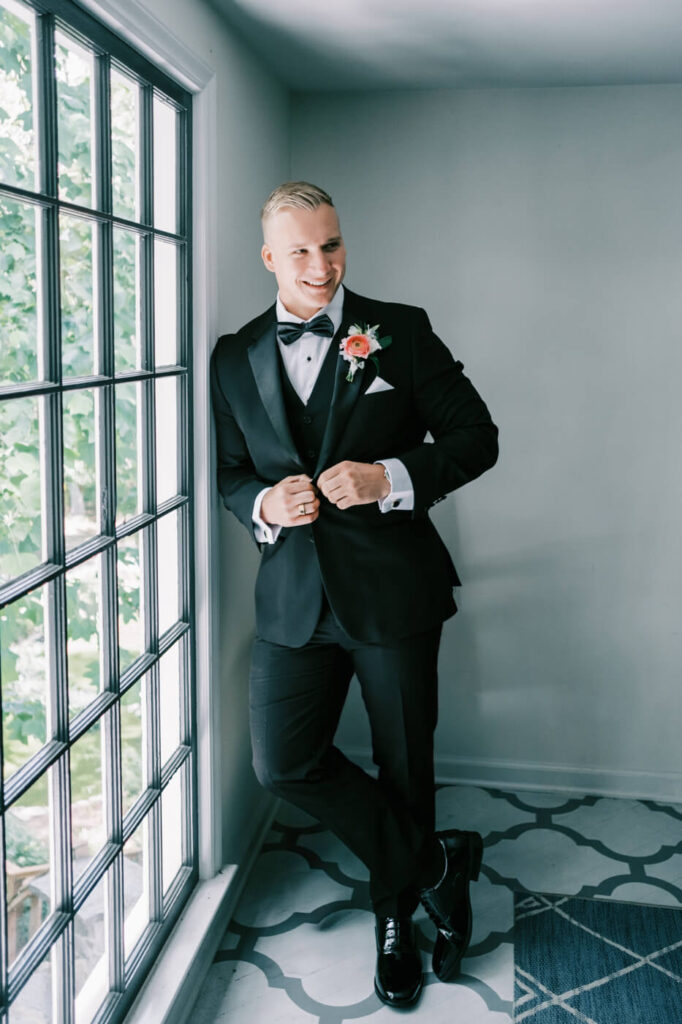 Groom adjusting his jacket with boutonniere while posing by a window at Historic Robertson House, looking sharp in a classic black tuxedo.