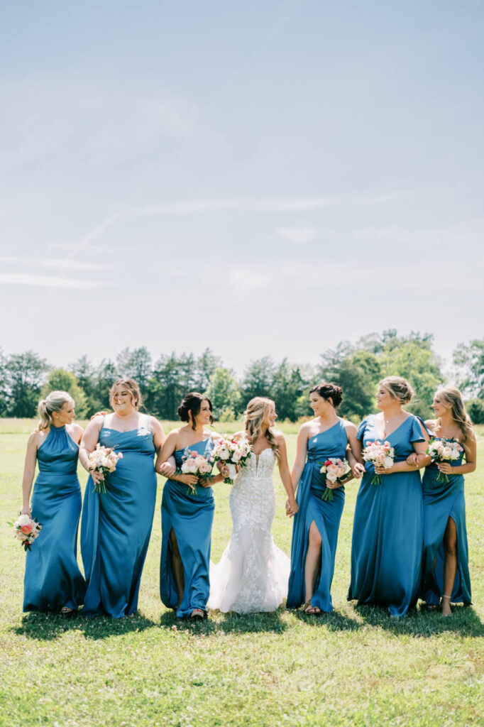 Bride walking with bridesmaids in blue dresses at The Historic Robertson House wedding – The bride and her bridesmaids stroll through the lush green venue, holding vibrant bouquets, showcasing the elegance of this timeless wedding.