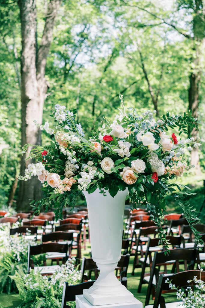 Grand floral arrangement at the outdoor wedding ceremony – A stunning white and blush floral centerpiece with lush greenery stands against a wooded backdrop, adding elegance to the wedding decor.