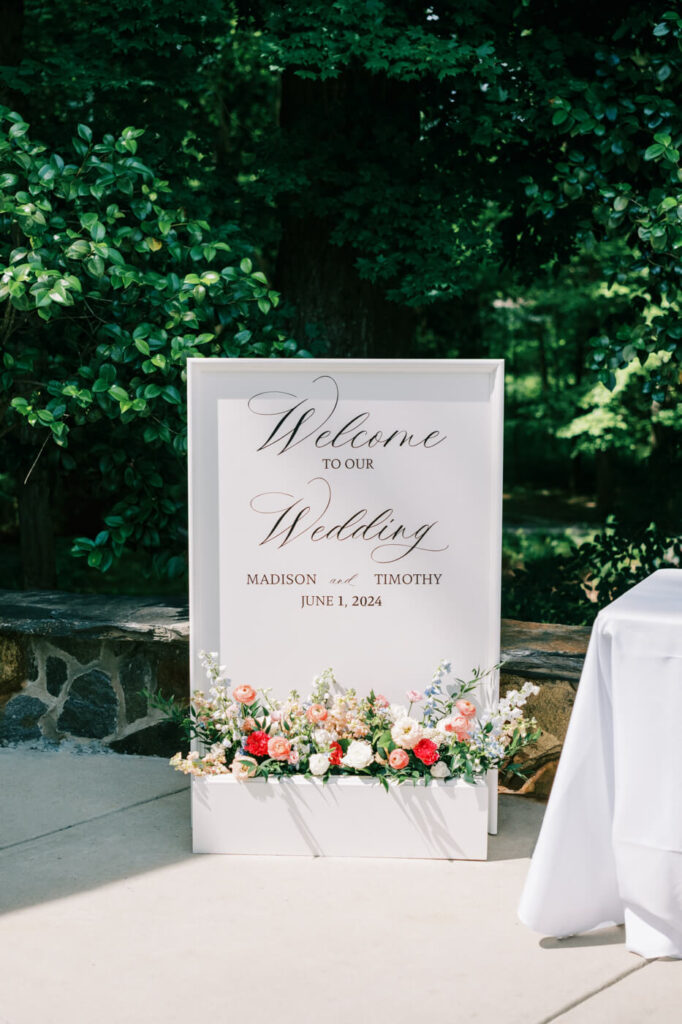 Elegant wedding welcome sign with floral arrangement – A beautifully designed white wedding welcome sign with cursive script and lush floral arrangements in coral, white, and blue hues at The Historic Robertson House.